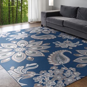 A blue area rug with large floral and paisley designs is placed in a modern living room with a gray sofa, a floor lamp, and a floor-to-ceiling window looking out to a green forest.