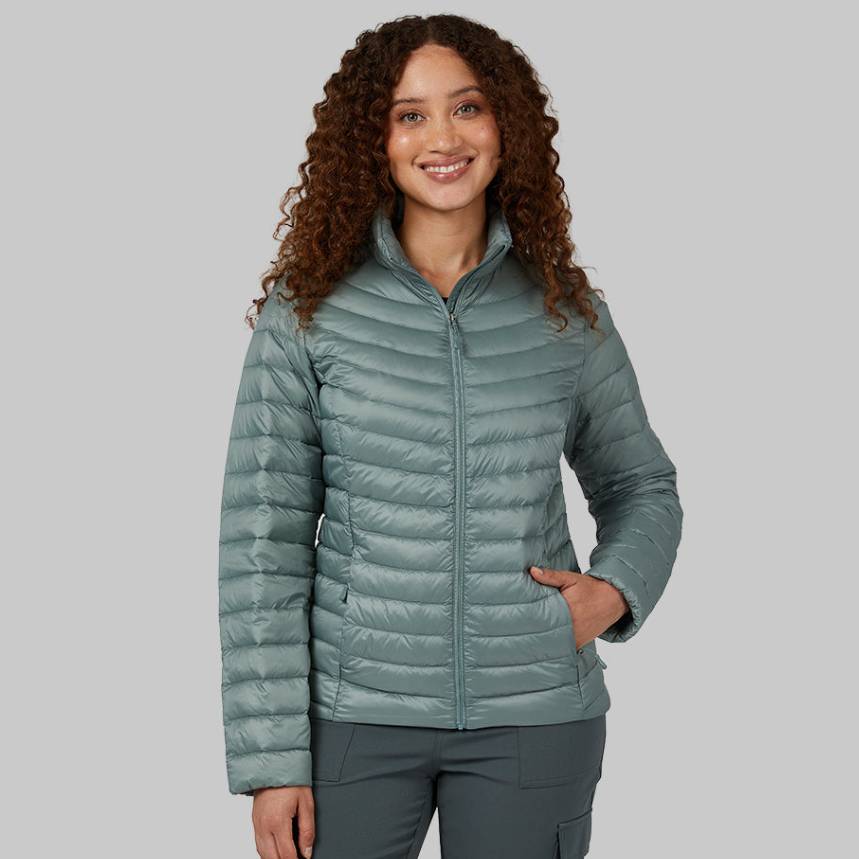 A woman is wearing a light green, quilted down jacket with a full-zip front and stand-up collar, designed to be lightweight and packable.