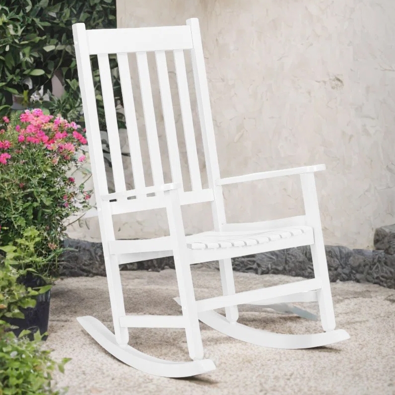 A white, solid wood rocking chair with a slatted back and seat, suitable for outdoor use. It's situated on a patio, surrounded by plants, with a textured stone wall background.