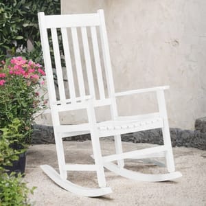 A white, solid wood rocking chair with a slatted back and seat, suitable for outdoor use. It's situated on a patio, surrounded by plants, with a textured stone wall background.