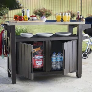 The Keter XL Outdoor Bar Cart is a mobile wood-like cart with a countertop, featuring storage space below for items like charcoal and bottled water. It has a side hook for tools and is set up with plates, cups of juice, and condiments.