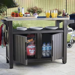 The Keter XL Outdoor Bar Cart is a mobile wood-like cart with a countertop, featuring storage space below for items like charcoal and bottled water. It has a side hook for tools and is set up with plates, cups of juice, and condiments.