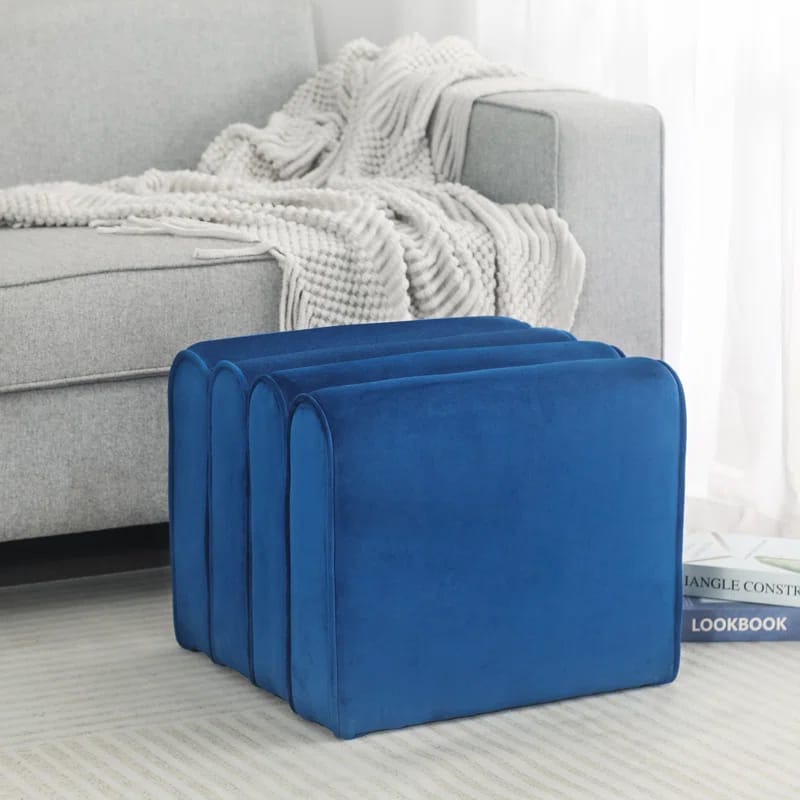 Blue velvet ottoman with a solid wood frame, featuring a modern, slightly curved design. It is positioned near a gray sofa adorned with a textured white blanket.