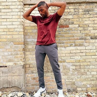 A man stands against a brick wall wearing a maroon T-shirt, gray joggers, and white sneakers.