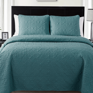A bed is made with a teal quilt featuring an embossed floral pattern, accompanied by matching pillowcases against a dark headboard.
