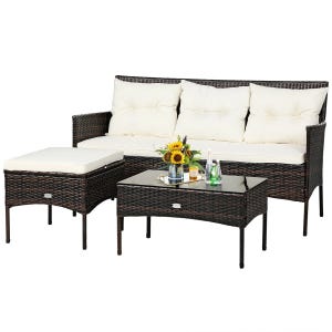 Outdoor patio set with a dark wicker three-seater sofa, cushioned ottoman, and a glass-top coffee table. The cushions are white, and a vase with sunflowers is placed on the table.