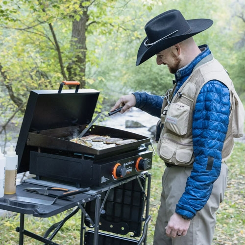 a man wearing a black cowboy hat grilling on a Blackstone griddle in the woods