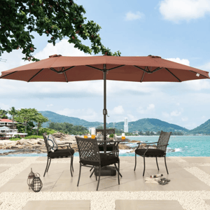 A patio setup with a large brown outdoor umbrella shading a round table and four chairs, overlooking a beach with mountains in the background.