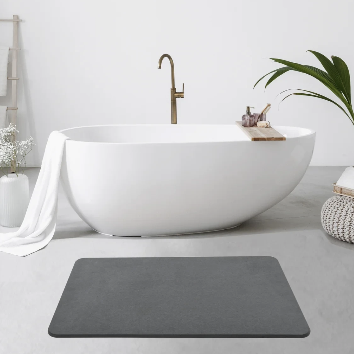 Modern bathroom with a white freestanding bathtub, brass faucet, wooden bathtub tray with toiletries, and a gray bath mat on the floor. Features a white towel, white vase with flowers, and a woven ottoman with a potted plant.