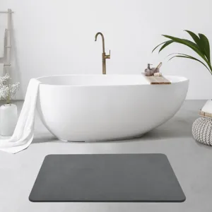 Modern bathroom with a white freestanding bathtub, brass faucet, wooden bathtub tray with toiletries, and a gray bath mat on the floor. Features a white towel, white vase with flowers, and a woven ottoman with a potted plant.