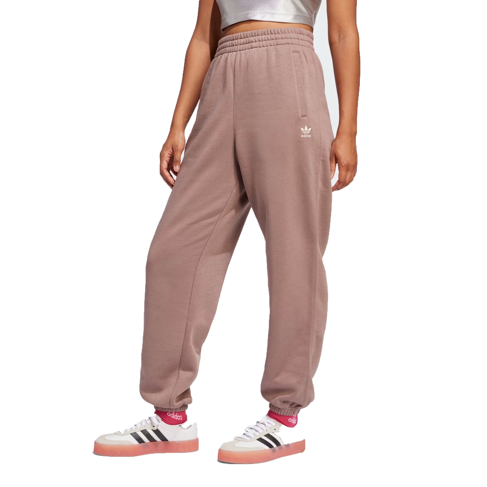 Adidas Essentials Loose Joggers are relaxed-fit, dusty pink sweatpants featuring an elastic waistband, side pockets, and the Adidas logo on the thigh. Paired with white Adidas shoes with black stripes and red accents.