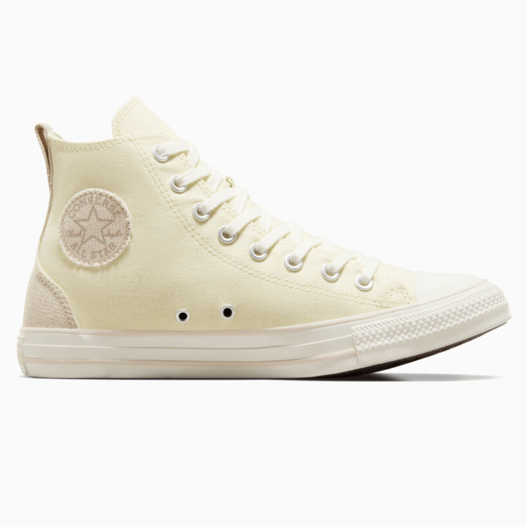 A high-top canvas sneaker in an off-white color with metal eyelets and a circular ankle patch logo.