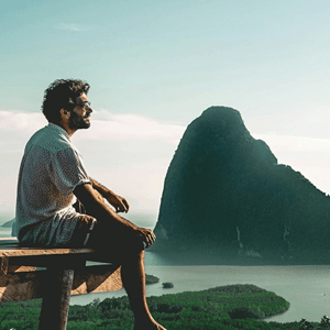 A person sits on a wooden platform overlooking a scenic, mountainous landscape with greenery and water below.