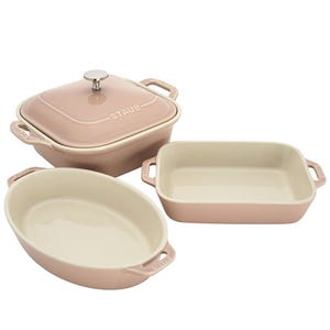 Three pink Staub ceramic baking dishes: one square with a lid, one oval, and one rectangular, all featuring side handles.