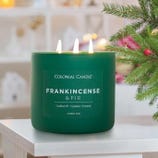 A green candle labeled \“Colonial Candle Frankincense & Fir\“ with notes of balsam fir, cypress, and incense, featuring three lit wicks and a festive setting with holiday decorations.