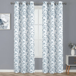 White curtains with a blue floral pattern hang on a rod in front of a window, against a gray wall. A small painting and a side table with a vase of flowers are beside them.