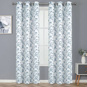 White curtains with a blue floral pattern hang on a rod in front of a window, against a gray wall. A small painting and a side table with a vase of flowers are beside them.