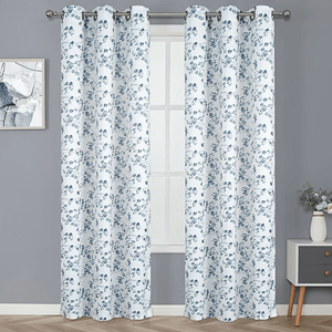 White curtains with a blue floral pattern hang on a rod in front of a window, against a gray wall. A small painting and a side table with a vase of flowers are beside them.