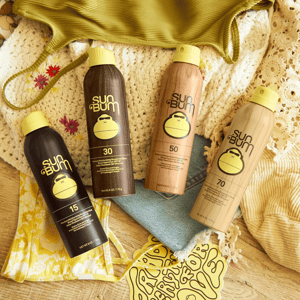 Four Sun Bum sunscreen spray bottles with SPF 15, 30, 50, and 70 are displayed on a wooden surface, accompanied by summer-themed clothing and accessories.