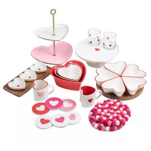 Heart-themed home decor set including heart-shaped plates, nesting bowls, a tiered tray, mugs, coasters, glasses, and a colorful trivet. Predominantly in red, pink, and white, featuring heart motifs.