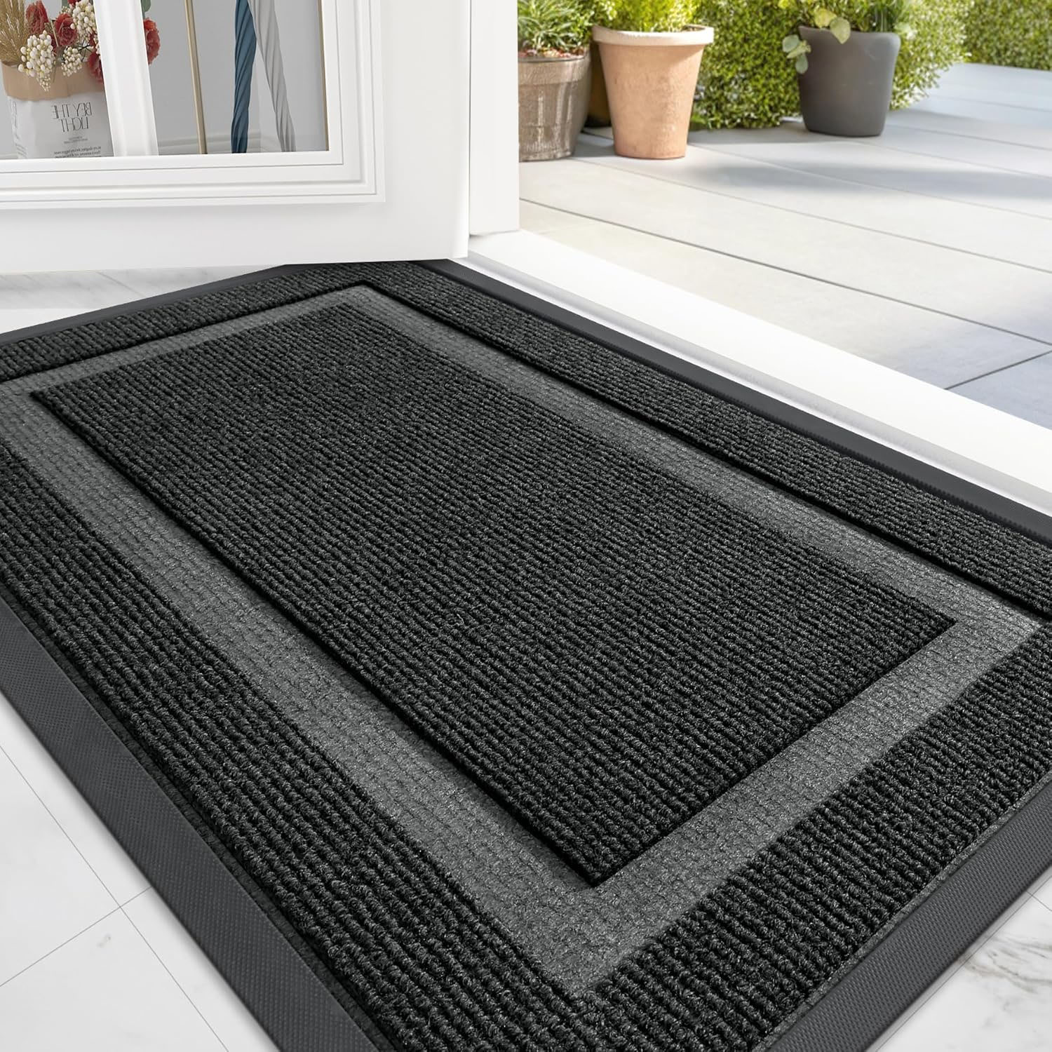Black textured doormat placed at a doorway with visible outdoor pots and plants.
