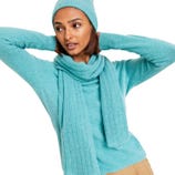 A woman wears a matching turquoise knit beanie, scarf, and sweater, paired with beige pants.