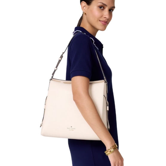 A woman carries a cream-colored Kate Spade Dumpling Shoulder Bag with a structured design, adjustable shoulder strap, and subtle logo detail, paired with a dark navy dress and gold bracelets.