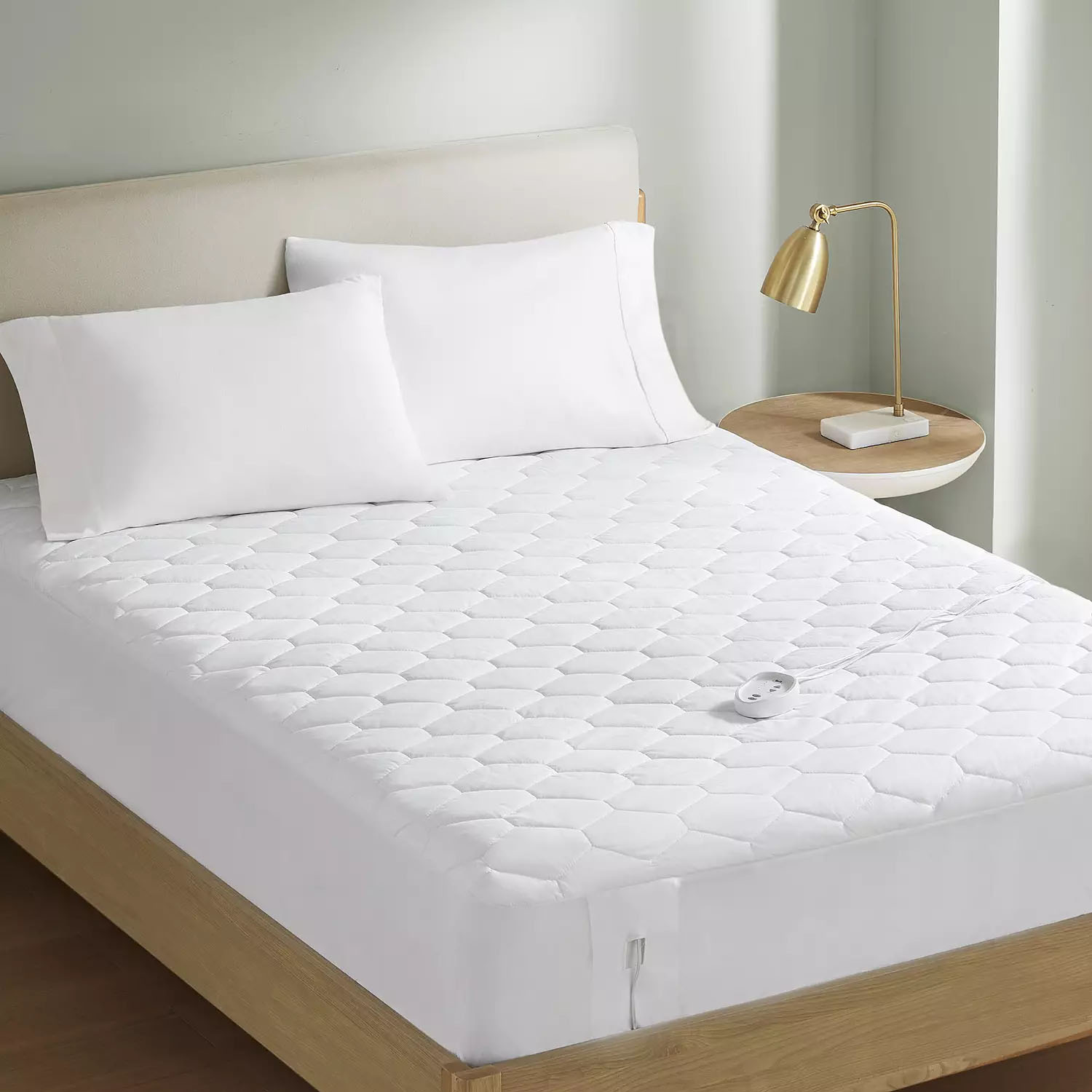 Serta Heated Mattress Pad features a quilted white design with a controller, resting on a bed with white pillows. A side table with a gold lamp accompanies the setup, offering a cozy and warm sleeping environment.