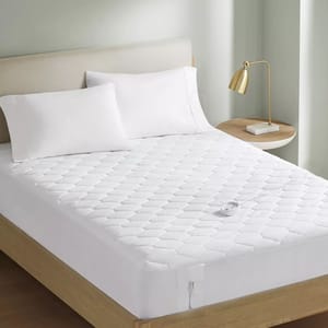 Serta Heated Mattress Pad features a quilted white design with a controller, resting on a bed with white pillows. A side table with a gold lamp accompanies the setup, offering a cozy and warm sleeping environment.