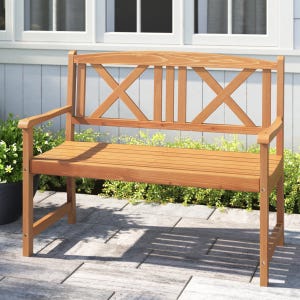 A wooden outdoor bench with a natural finish, featuring a slatted seat and decorative X-back design, set on a stone patio next to greenery.