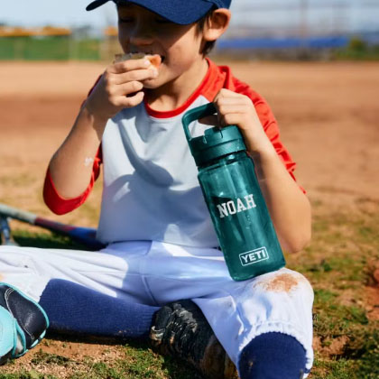 A young child wearing a baseball uniform sits on a field, holding a YETI water bottle personalized with the name \“Noah.\“