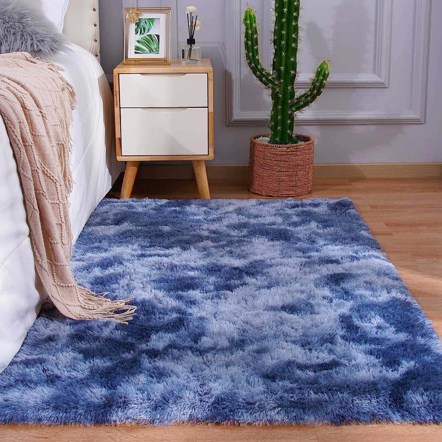 A blue and white fluffy rug is placed on a wooden floor next to a bed with a beige blanket. A small wooden nightstand with drawers holds decor items, next to a cactus in a woven basket.