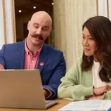 Two people are seated at a table looking at a laptop. The man gestures toward the screen, while paperwork is spread out on the table next to the woman.
