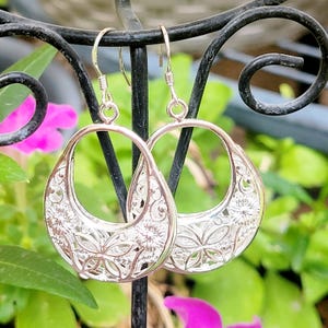 Dangling earrings with intricate floral and leaf cut-out designs hanging from a decorative metal stand, set against a backdrop of blurred green leaves and pink flowers.