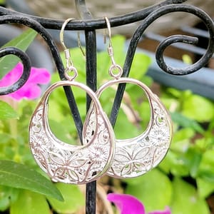Dangling earrings with intricate floral and leaf cut-out designs hanging from a decorative metal stand, set against a backdrop of blurred green leaves and pink flowers.