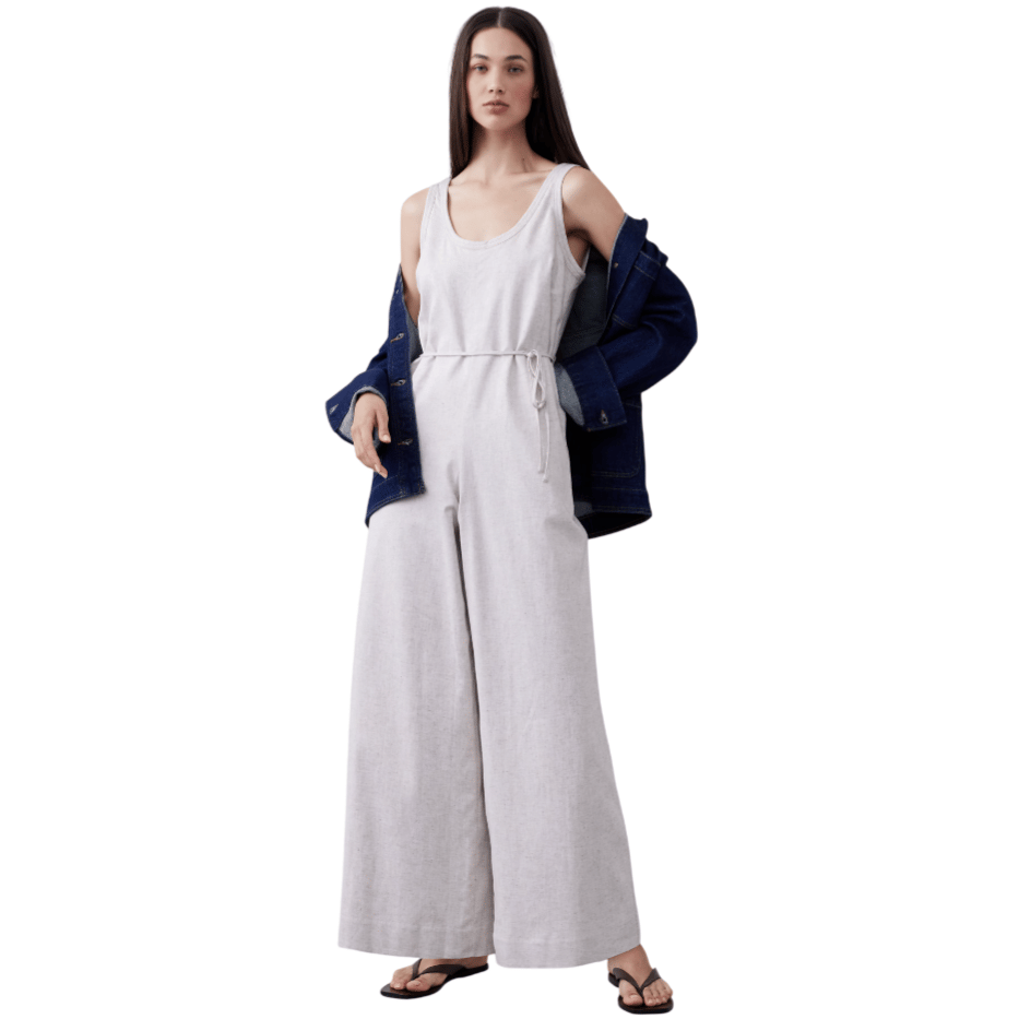 A woman wears a light-colored sleeveless jumpsuit with a tie belt, paired with a dark denim jacket and black sandals, standing against a minimalistic background.