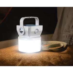 A portable LED lantern with a handle and flashlight feature, emitting bright light and placed on a surface. A strap is positioned nearby for carrying.