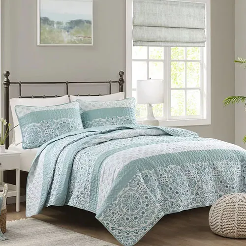 A bed is shown with light blue and white patterned bedding, including a quilt and matching pillow shams, in a bright room featuring a white lamp, green plant, and framed wall art.