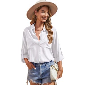 A woman wears a white long-sleeve button-up shirt, denim shorts, a wide-brimmed beige hat, large hoop earrings, and a chain necklace. She carries a small quilted white handbag with a gold chain strap.