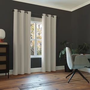 Light gray blackout curtains hang on a dark rod in front of a window with white trim, set against a dark wall. The room features wood flooring, a modern gray chair, a small dresser, and a houseplant.
