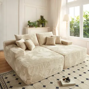 A plush, cream-colored sectional sofa with ribbed texture and several matching cushions sits in a bright living room with large windows, a patterned rug, and potted plants on a mantel.