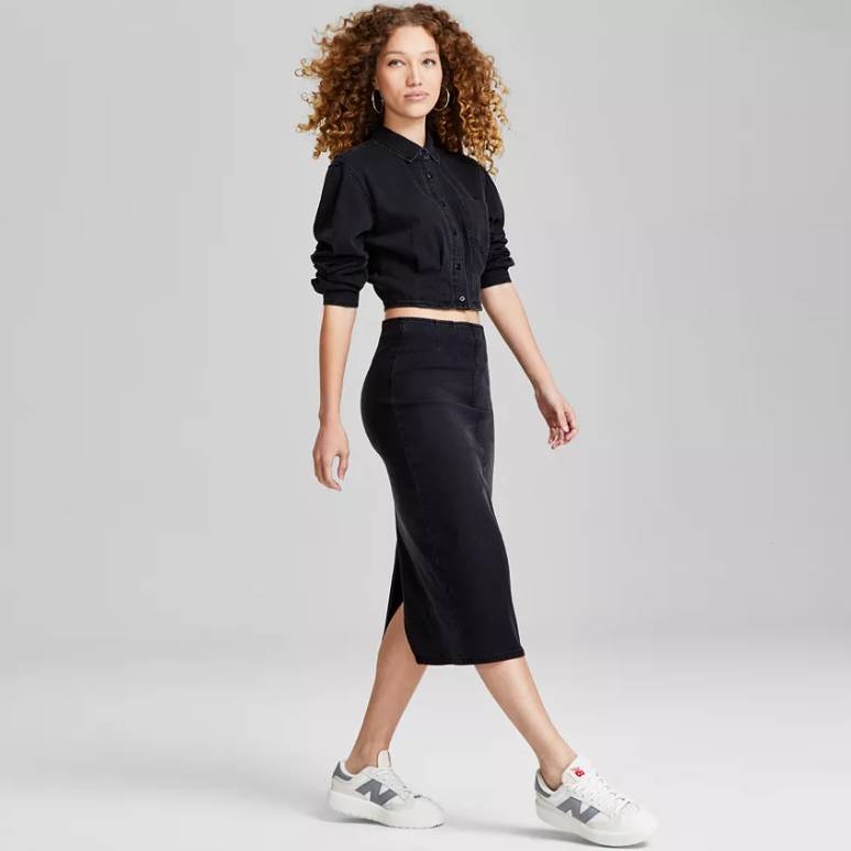 A woman models a high-waisted, black denim midi skirt with a back slit, paired with a black button-up shirt and white sneakers.