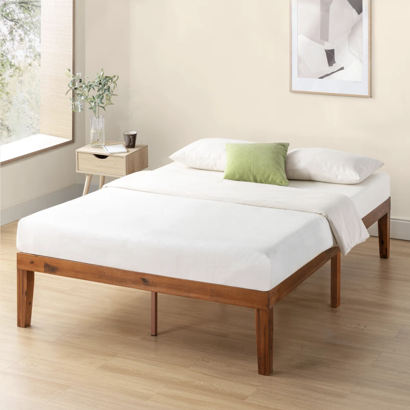 A minimalist wooden bed frame with a white mattress, accompanied by two white pillows and a green accent pillow, next to a wooden nightstand with a plant and a cup.