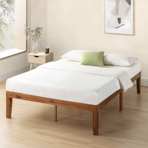 A minimalist wooden bed frame with a white mattress, accompanied by two white pillows and a green accent pillow, next to a wooden nightstand with a plant and a cup.