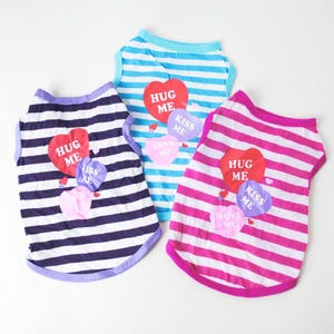 Three striped dog shirts in blue, purple, and pink, each with a heart-shaped sticker saying \“Hug Me,\“ \“Kiss Me,\“ and \“Love Me.\“