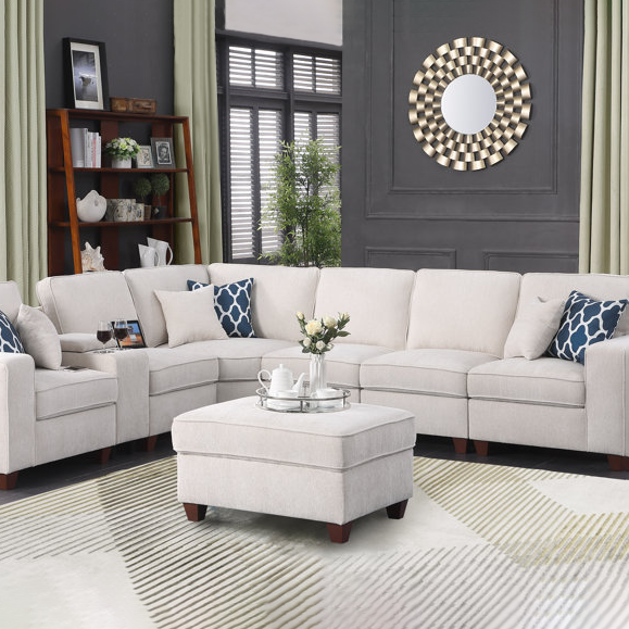 A beige sectional sofa with matching ottoman, two blue and white patterned throw pillows, and a decorative round mirror on the wall. A wooden bookshelf with decorative items sits in the corner, and a light-colored rug covers the floor.