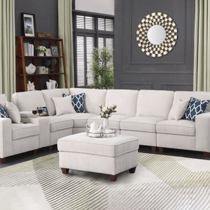 A beige sectional sofa with matching ottoman, two blue and white patterned throw pillows, and a decorative round mirror on the wall. A wooden bookshelf with decorative items sits in the corner, and a light-colored rug covers the floor.