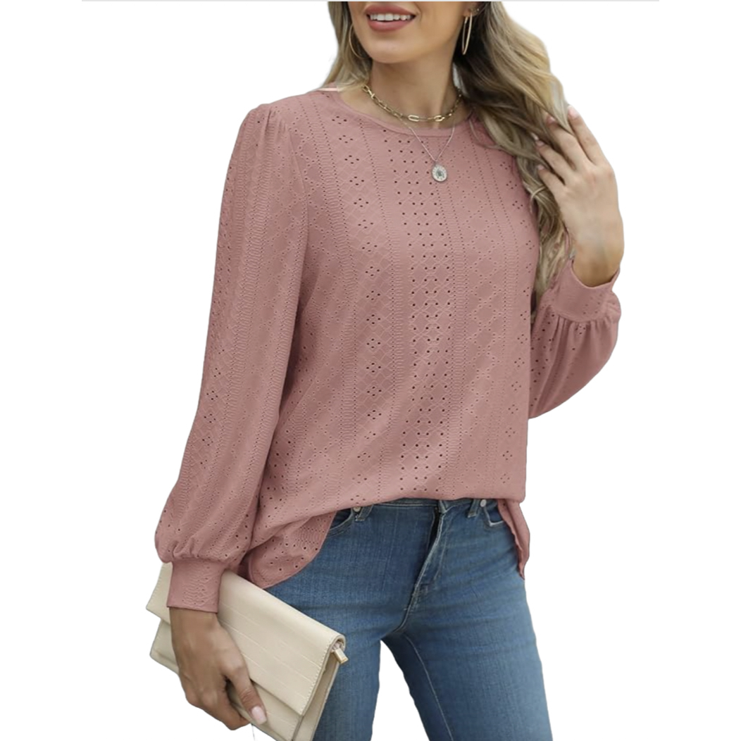 A woman wears a pink eyelet blouse with long sleeves and a round neckline, paired with blue jeans. She holds a beige clutch and wears a necklace and hoop earrings.