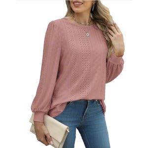 A woman wears a pink eyelet blouse with long sleeves and a round neckline, paired with blue jeans. She holds a beige clutch and wears a necklace and hoop earrings.