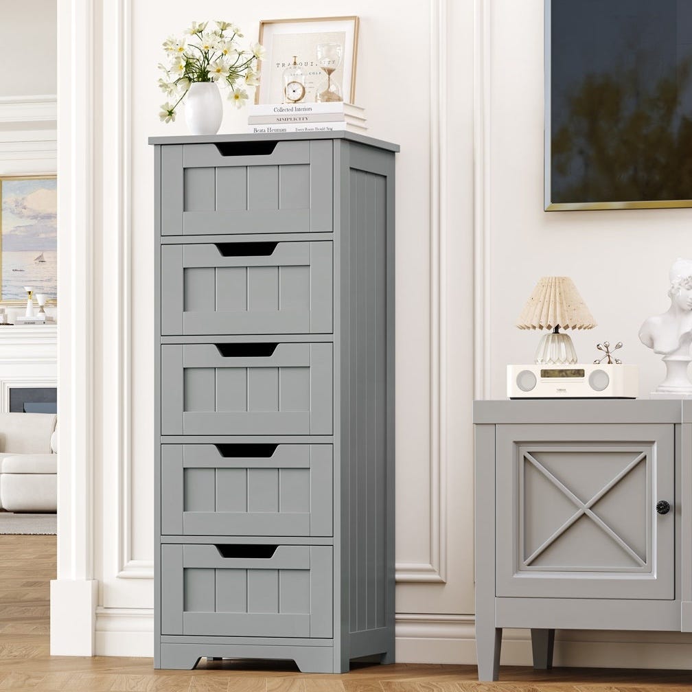 Tall gray dresser with five drawers, featuring a classic panel design, topped with decorative items like a vase, books, and framed pictures. Adjacent is a smaller cabinet with a decorative lamp and bust.